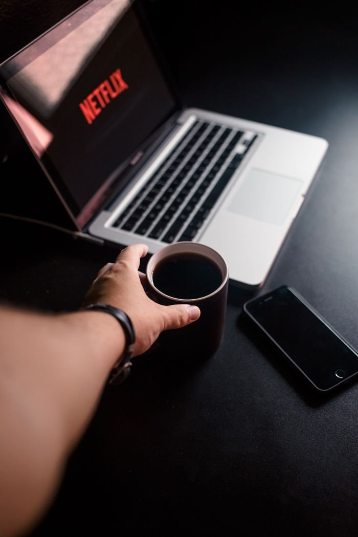 A hand reaching for coffee by a laptop streaming Netflix. Cozy, tech-infused leisure moment.