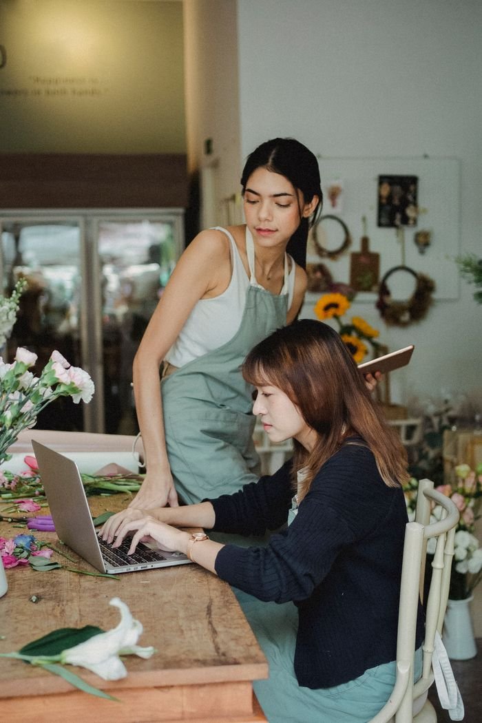 Focused multiethnic female florists sitting at table and typing on computer while checking online orders during work in floral store